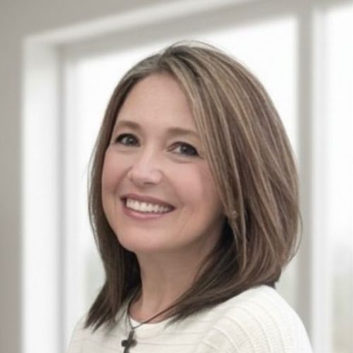 A woman with shoulder-length brown hair and blonde highlights smiles at the camera. She is wearing a white sweater and a necklace with a small cross pendant, standing in front of large, bright windows.